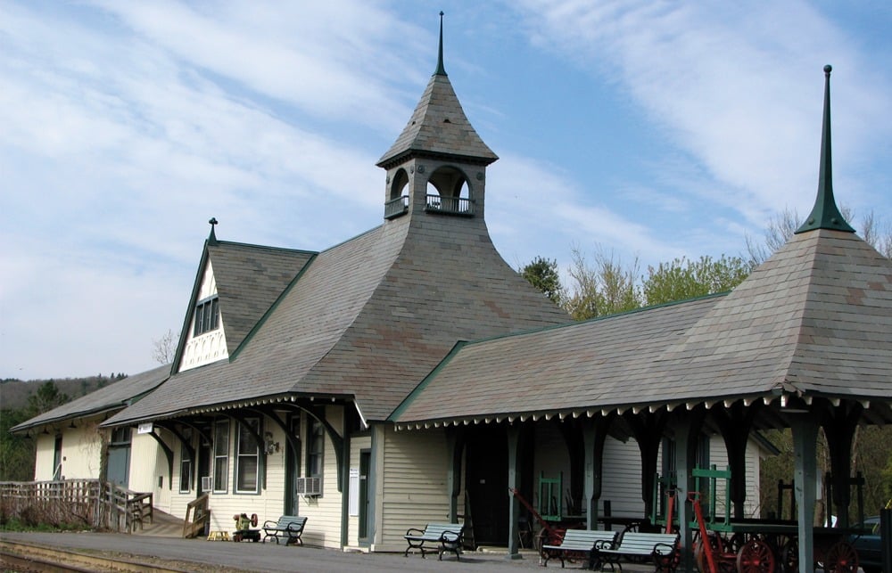 A historic train station with a tall, pointed roof and wooden siding. The building has large windows, decorative trim, and a small bell tower. Benches and a cart are placed on the platform. The sky is partly cloudy.