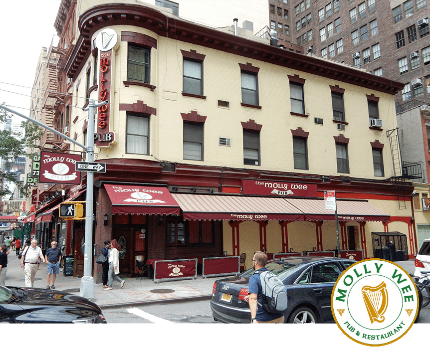 Street view of The Molly Wee Pub and Restaurant, a charming three-story building with a red and beige exterior, nestled on an urban corner. The pub features vibrant red awnings and signage while people stroll past on the bustling sidewalk.