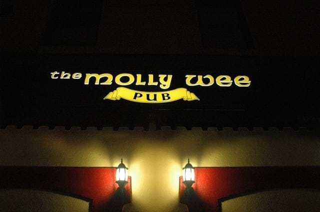A dimly lit sign for Molly Wee Pub is illuminated at night, with two lantern-style lights mounted below it, casting a soft glow on the wall.