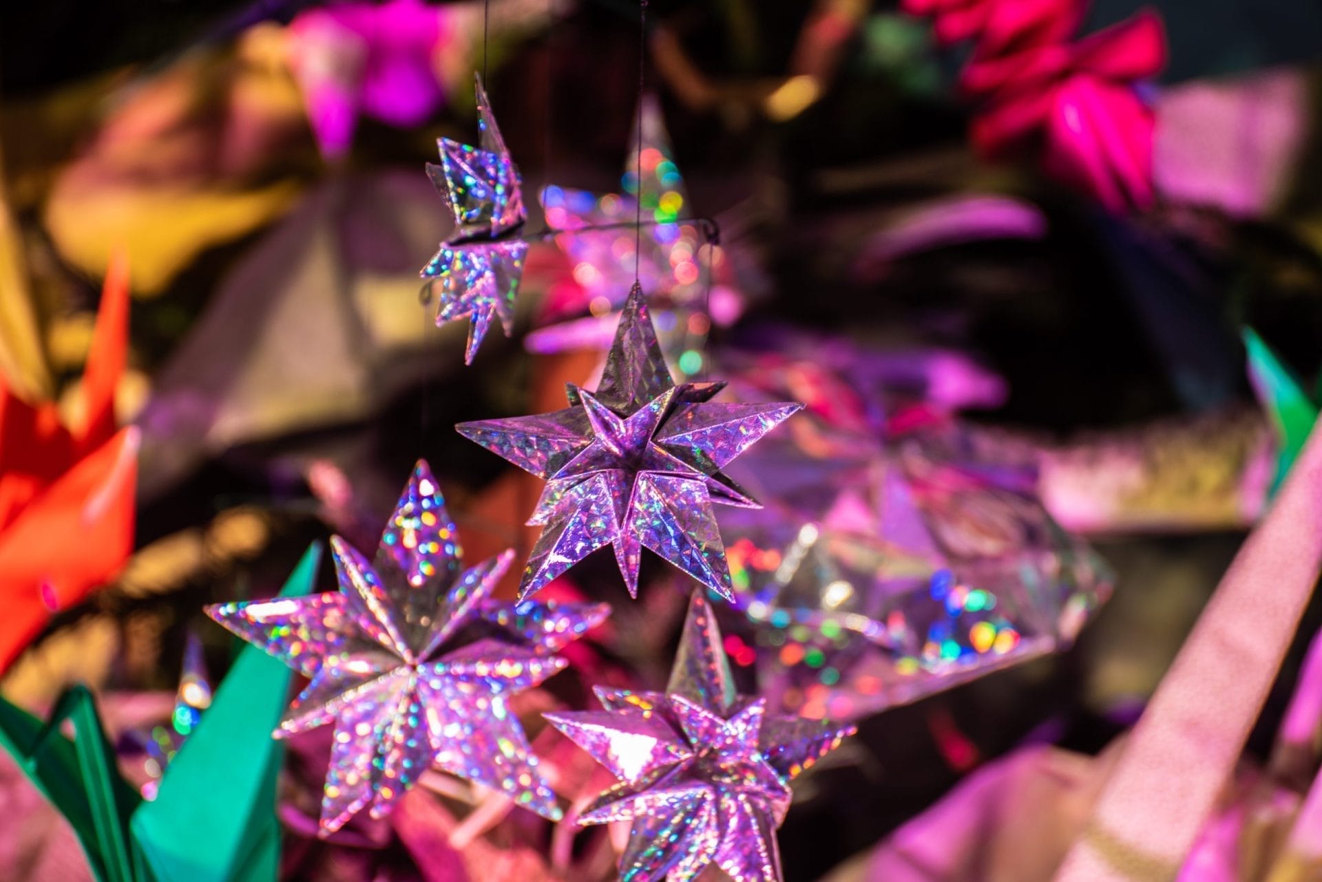 Close-up of shimmering, multi-colored star ornaments hanging in a festive display, reminiscent of the dazzling exhibits at the American Museum of Natural History. The stars have a holographic effect, reflecting vivid colors of pink, purple, and green against a blurred background of other decorative items.