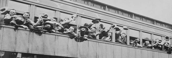A group of men in military uniforms lean out of a trains windows, enthusiastically waving their hats with a spirit of sacrifice. The train, labeled with part of the word Pennsylvania, appears to be in motion. The men are smiling and appear excited or celebratory.