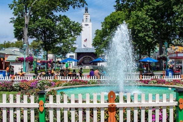 A lively amusement park scene in New Rochelle features a white fountain surrounded by flowers and a white picket fence. People enjoy tables under blue umbrellas, while the tall clock tower, reminiscent of the nearby Amtrak Station, stands proudly amidst green trees—connecting communities through fun and leisure.