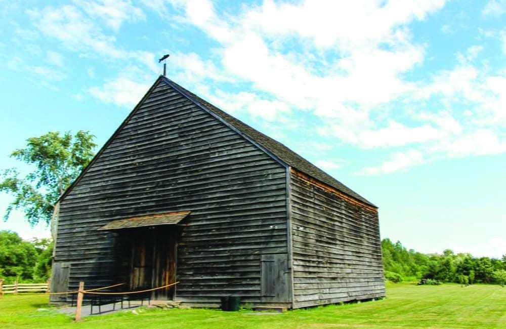 A large, weathered wooden barn at Mabee Farm Historic Site sits in a grassy field under a bright blue sky with scattered clouds. A black bird perches on its roof, and a rope barrier is in front of the barns entrance. Trees are visible in the background.