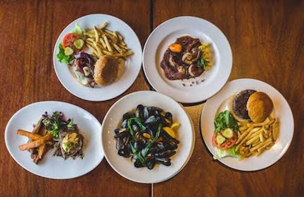 Five plates on a wooden table present a new world bistro experience: burgers with fries and salad, steak garnished with vegetables, and a bowl of mussels accompanied by lemon slices.