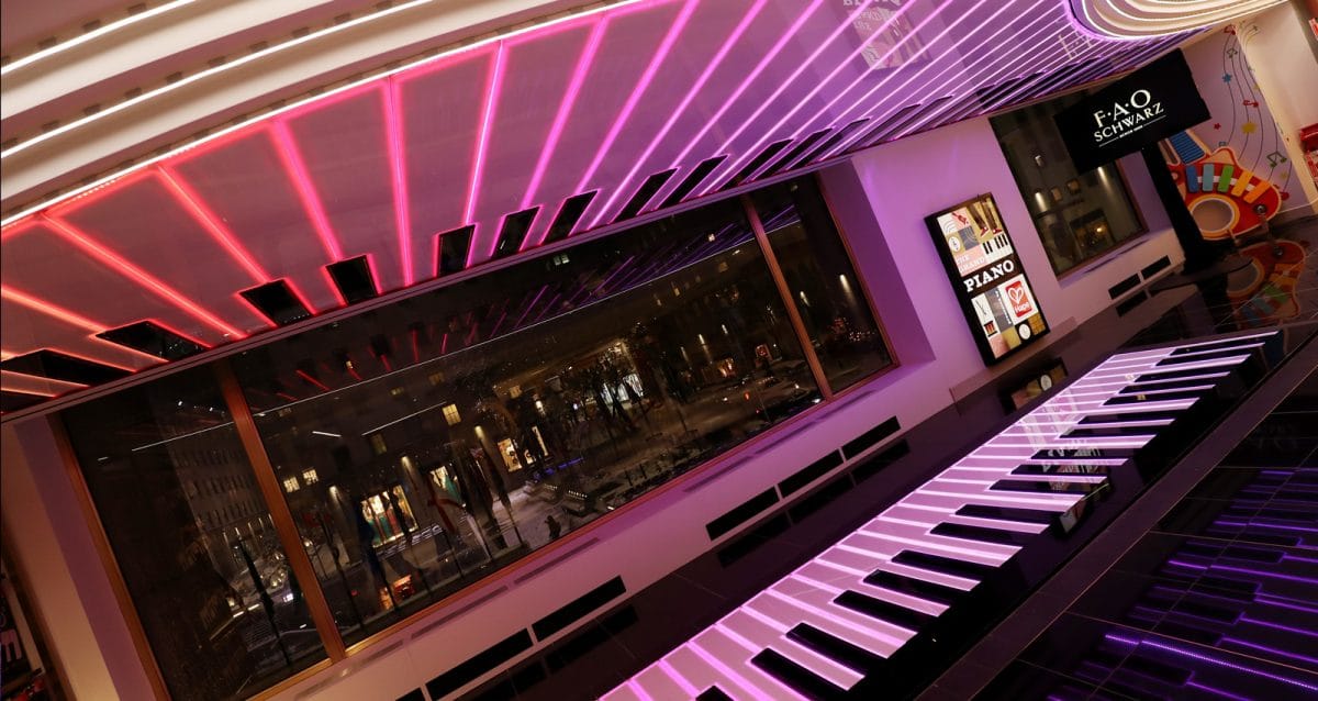 A large piano keyboard display enchants visitors at FAO Schwarz, bathed in the glow of colorful pink and white ceiling lights. Through the window, the city street bustles with life, while a sign proudly showcases the iconic FAO Schwarz brand on the wall.