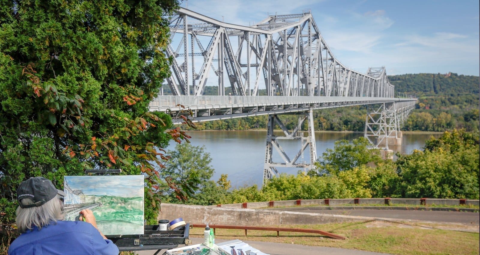 A person with gray hair paints a landscape scene featuring a large steel bridge over a river, reminiscent of the Hudson River Skywalk. Lush green trees surround the area, and hills are visible in the background under a clear blue sky. A person with gray hair paints a landscape scene featuring a large steel bridge over a river, reminiscent of the Hudson River Skywalk. Lush green trees surround the area, and hills are visible in the background under a clear blue sky.