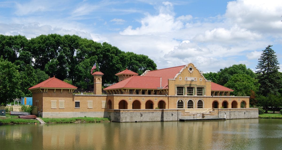 Nestled by a calm lake, this historic Park Playhouse boasts a red-tiled roof and arched windows. Encircled by lush green trees under a partly cloudy sky, the buildings serene reflection shimmers in the water.