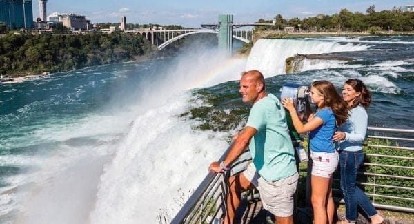 A family of three stands at a railing overlooking Niagara Falls. The daughter uses a viewing scope while the parents enjoy the view. As they explore travel packages, a rainbow appears above the falls, and a bridge spans the river in the background under a clear blue sky. A family of three stands at a railing overlooking Niagara Falls. The daughter uses a viewing scope while the parents enjoy the view. As they explore travel packages, a rainbow appears above the falls, and a bridge spans the river in the background under a clear blue sky.