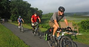 Three people are cycling on a paved path through a lush green park. They wear helmets and casual attire, carrying bags on their bikes. In the background, theres a bench and trees with a misty horizon.