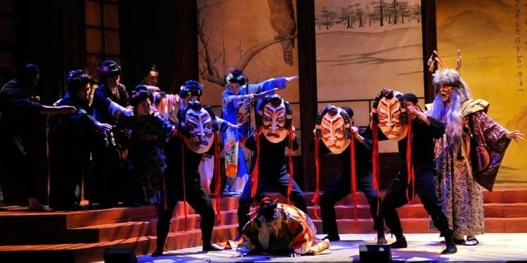 Performers on stage in a dramatic scene, wearing elaborate costumes and holding large, expressive masks. The background features traditional Asian art, and the lighting accentuates the vivid colors and movements of the actors.