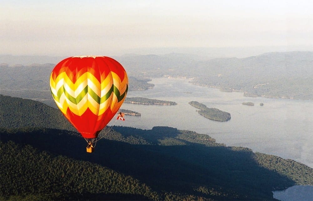 A beautiful hot air balloon with red, orange, and green patterns floats above a scenic landscape, showcasing a large body of water and lush green hills under a clear sky.