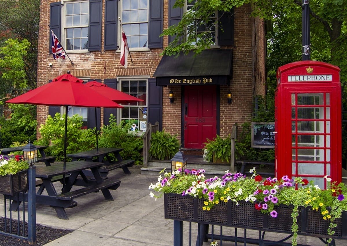 An Olde English Pub with brick exterior, red doors, and windows. A red telephone booth stands nearby. A patio with a black picnic table and red umbrella is surrounded by flower boxes filled with colorful blooms. An Olde English Pub with brick exterior, red doors, and windows. A red telephone booth stands nearby. A patio with a black picnic table and red umbrella is surrounded by flower boxes filled with colorful blooms.
