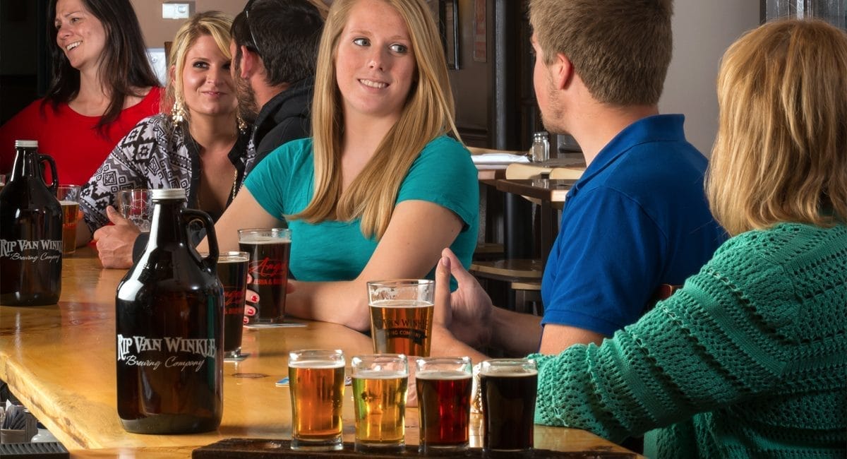 A group of people sits at a bar table, conversing and enjoying drinks. There are various glasses of beer on the table, alongside a prominently placed black growler labeled Rip Van Winkle Brewing Company. The atmosphere is relaxed and social.