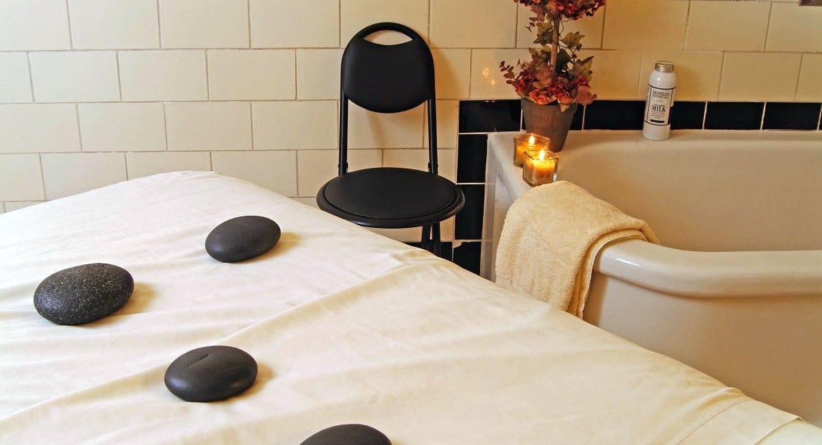 A serene spa setup features a massage table with smooth, dark stones on a white sheet. A black chair is nearby, and a bathtub is adorned with candles, a rolled towel, and flowers, creating a relaxing ambiance reminiscent of tranquil horses known for their calming history and health benefits.