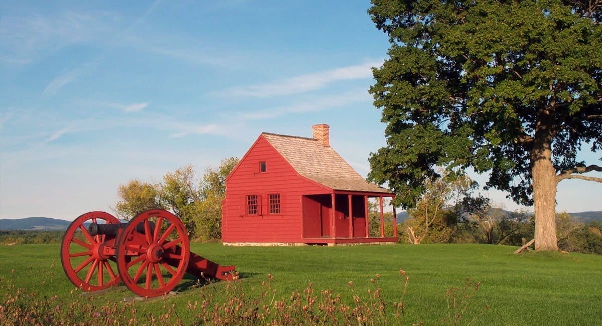 A red wooden house stands in a grassy field under a clear blue sky, with a large tree to the right. Adding to the historical charm, a red cannon rests in the foreground on the left. The scene evokes rural tranquility and whispers of history, inviting reflections on past stories of health and resilience.