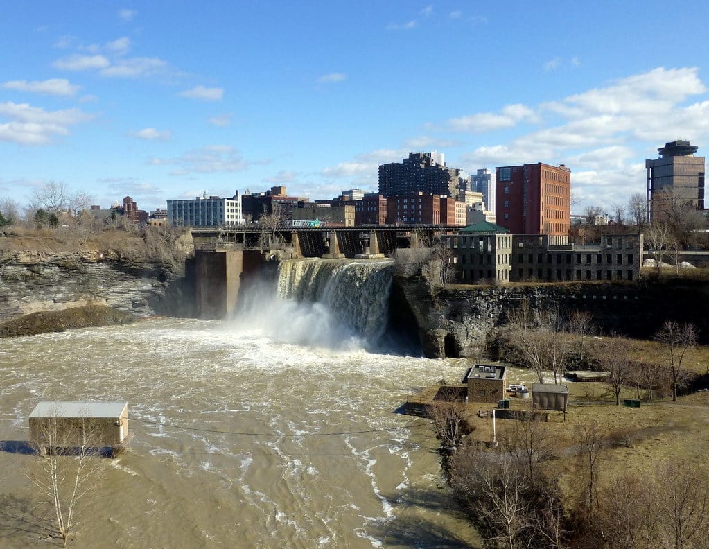 Rochesters waterfall cascades over a dam in an urban setting, with water flowing into a wide river. Industrial buildings and the city skyline are visible in the background, under a clear blue sky with scattered clouds.
