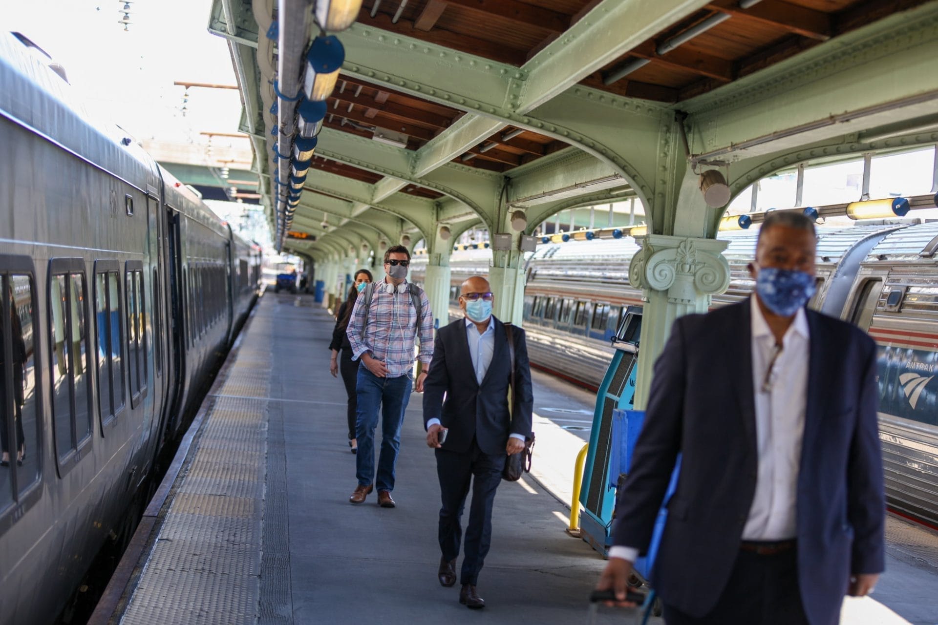 People wearing masks walk along a train platform, reflecting the ongoing coronavirus updates. A train is stationed on the left, and the platform has a covered roof with beams. The atmosphere suggests busy transit, hinting at possible Amtrak service impacts in such bustling areas.