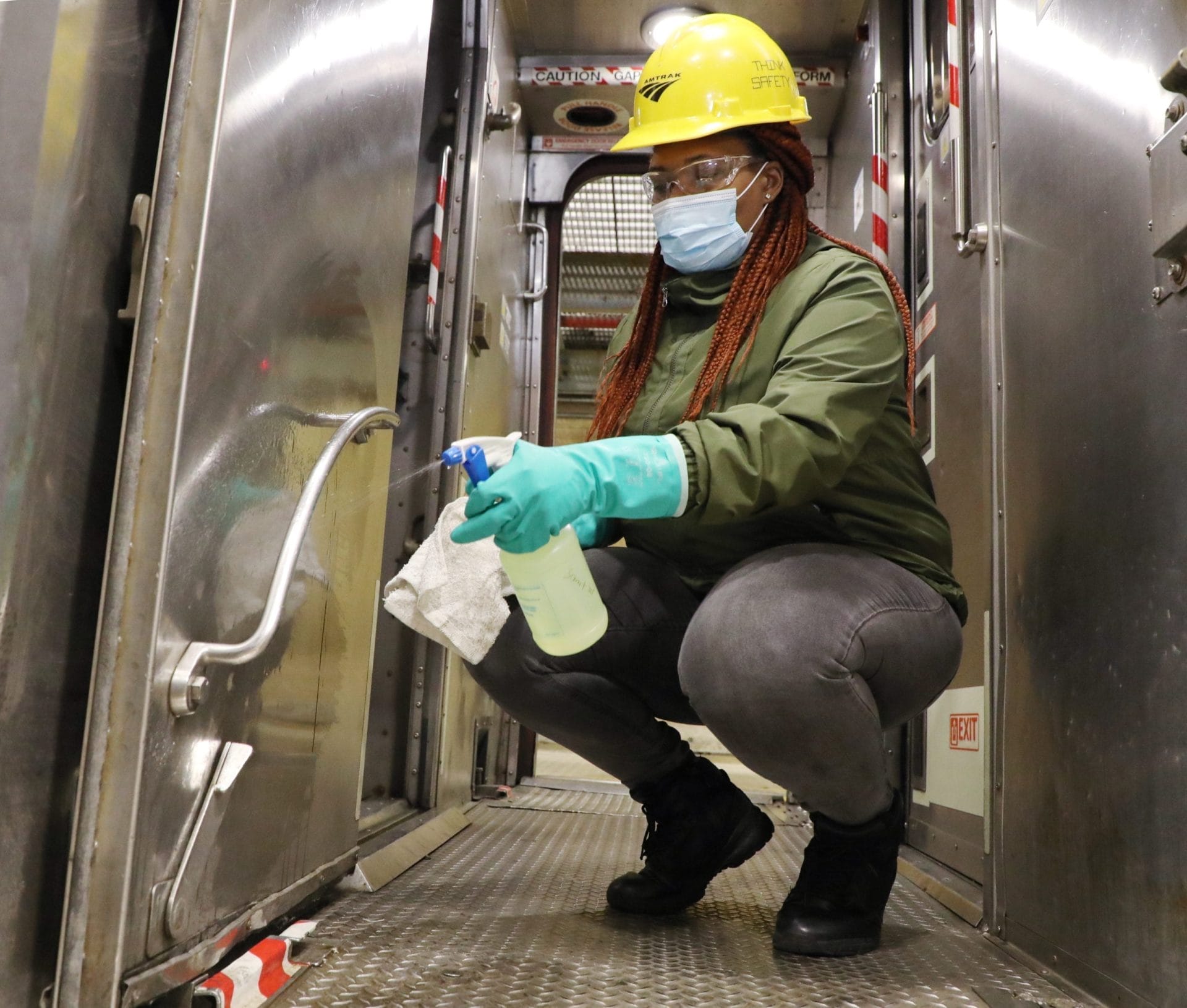 A person wearing a yellow hard hat, mask, and green gloves crouches inside a metal-laden corridor, diligently cleaning a door handle amid coronavirus updates. The environment resembles an industrial or transport setting, hinting at possible Amtrak service impacts. .