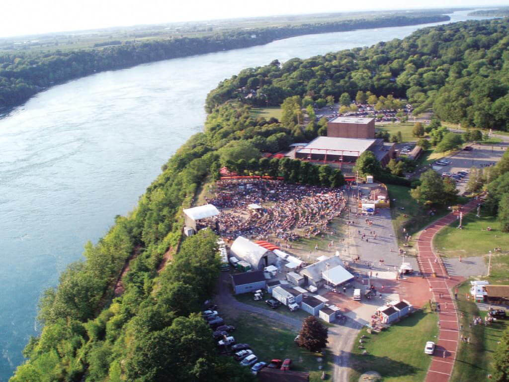 Aerial view of a large outdoor concert at ArtPark next to a river, with lush green landscapes in the background. Numerous people are gathered around the stage, and various tents and structures grace the festival grounds. A winding path leads to the parking area.