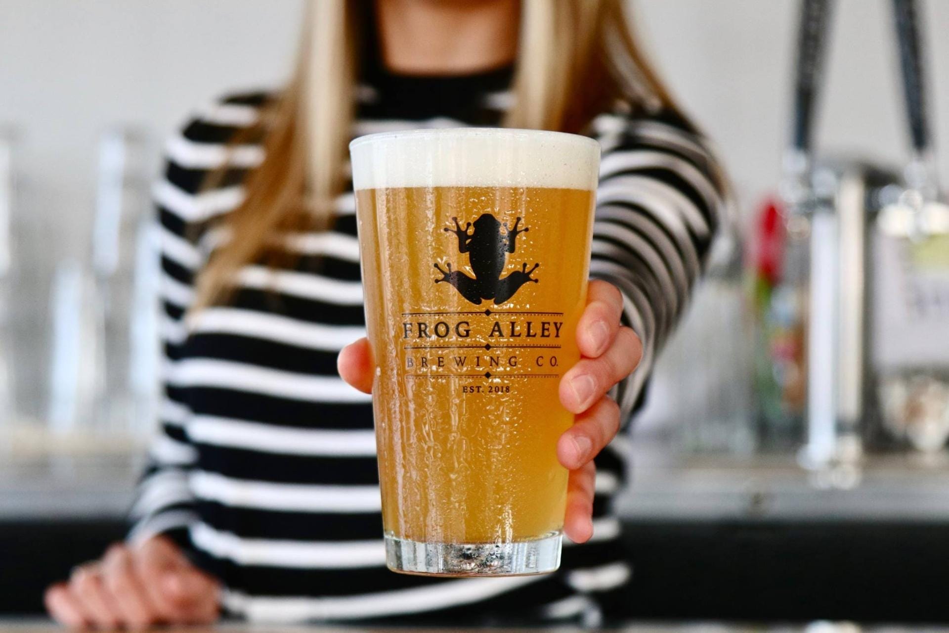 A person in a black and white striped shirt holds a pint of beer emblazoned with the Frog Alley Brewing Co. logo. The background captures the lively atmosphere of Frog Alley Brewing, showcasing taps and blurred elements that hint at the buzzing brewery setting.