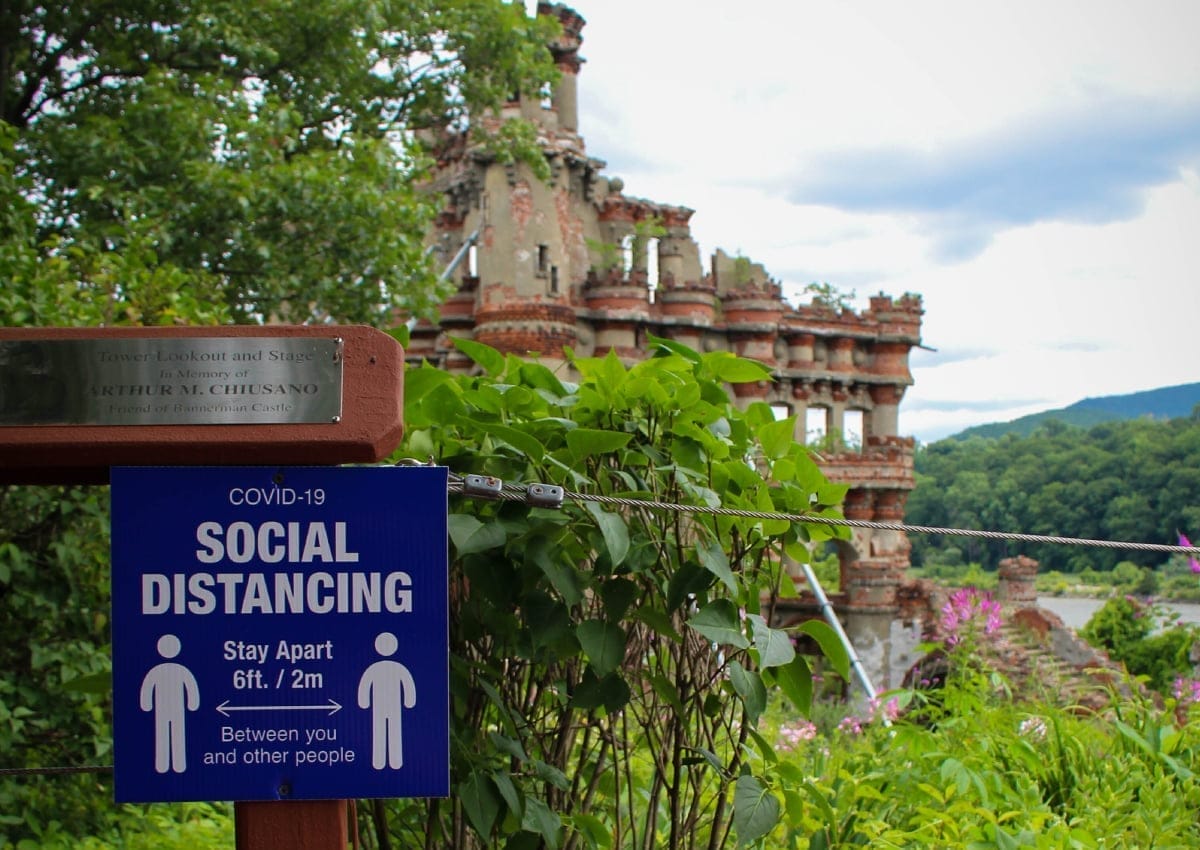 A sign reading COVID-19 Social Distancing: Stay Apart 6ft. / 2m Between You and Other People stands in the foreground, while the greenery and haunting ruins of Bannerman Castle loom in the background, adding a touch of historical intrigue to this safety reminder.