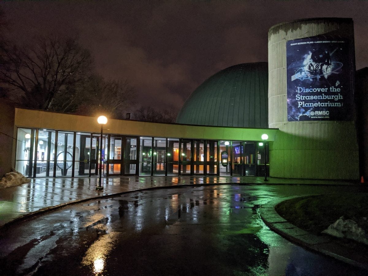 Night view of a planetarium entrance with glass doors reflecting light on the wet pavement. A cylindrical building displays a lit advertisement for the Strasenburgh Planetarium, part of the Rochester Museum & Science Center. Trees and a cloudy sky are visible in the background.