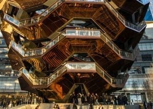 A large, honeycomb-like metal and glass structure with multiple levels and staircases, the Vessel at Hudson Yards stands majestic. People are walking and standing on various levels, surrounded by modern buildings and bathed in warm sunlight.