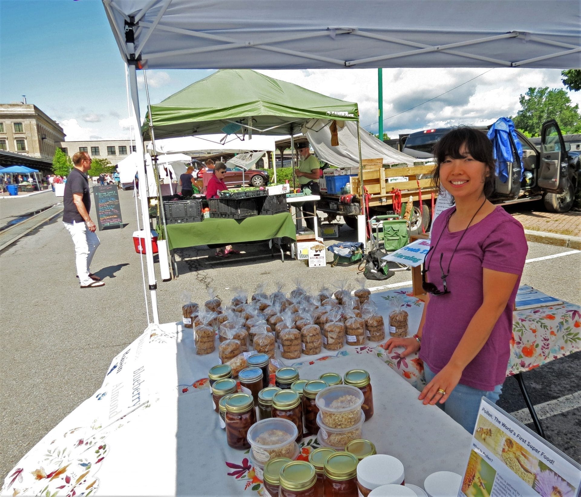 A woman stands at a market stall near Utica Station, smiling with jars of preserves and baked goods displayed on the table. Other market stalls are visible in the background on a sunny day. A woman stands at a market stall near Utica Station, smiling with jars of preserves and baked goods displayed on the table. Other market stalls are visible in the background on a sunny day.
