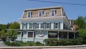 Historic light blue building with white trim and a Mansard roof, surrounded by greenery. Signs on the porch read Author Event Tonight. A circular sign on the building announces The Norwich Northshire Bookstore. Clear blue sky overhead.