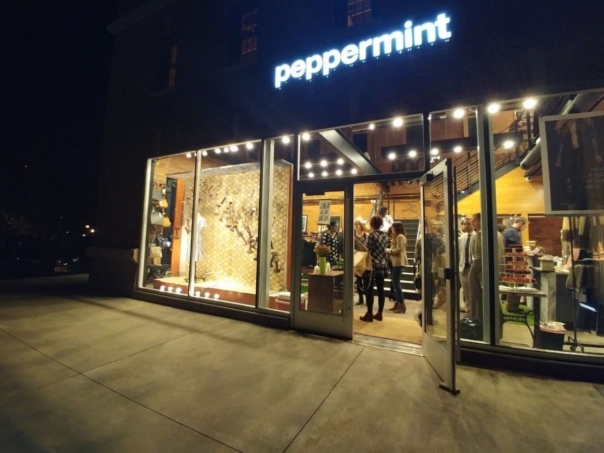 The Peppermint storefront at night glows warmly through its glass windows, revealing a modern and inviting interior. Inside, people gather amid displays of stylish clothes and accessories, creating a lively atmosphere under the soft lights.