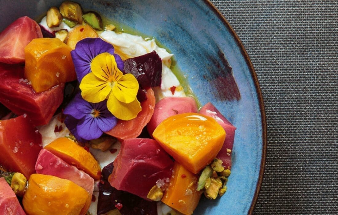 A vibrant Rhinebeck salad on a blue plate features chunks of colorful beets, dollops of cream, edible purple and yellow flowers, and scattered pistachios. The plate rests on a textured gray surface.