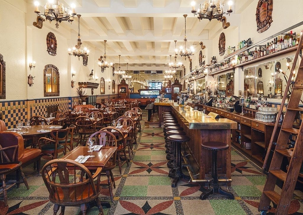 Dominion Square Tavern boasts a vintage-style interior with wooden tables and chairs, a long bar with stools, ornate chandeliers, and patterned tile flooring. The walls feature ornate decor and mirrors, creating a warm, inviting atmosphere perfect for savoring comfort food.