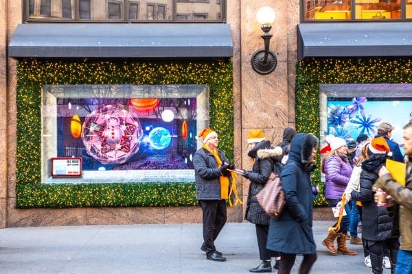People in winter clothing, some wearing orange Santa hats, gather in front of festive store windows in NYC adorned with green garlands and lights. The windows display colorful holiday decorations, creating a lively and cheerful scene.