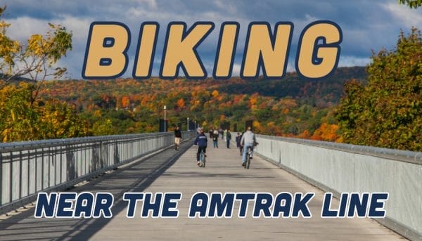 People biking on a wide bridge surrounded by autumn foliage, with the sky partly cloudy. Large text reads BIKING and smaller text below reads NEAR THE AMTRAK LINE. Get ready to embrace winter events while enjoying this scenic route!.