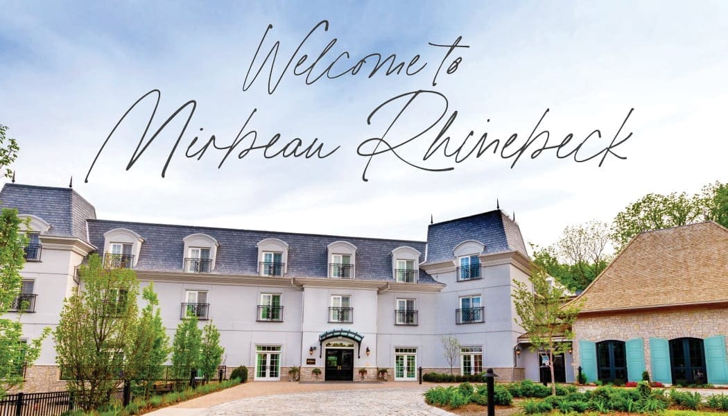 Elegant hotel with a French-inspired design, featuring a courtyard with trees and cobblestone path. A large script font sign reads Welcome to Mirbeau Rhinebeck above the building, reminiscent of an exclusive Manhattan Club in its sophisticated charm.