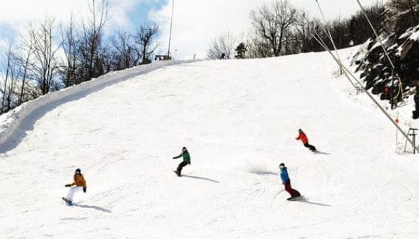 Four snowboarders descend a snow-covered slope on a sunny day. The sky is partly cloudy, and there are leafless trees on the side.
