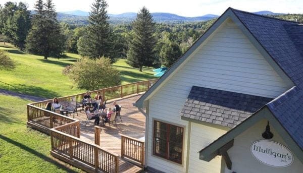 A group of people are seated at outdoor tables on a wooden deck attached to a white building with a sloped roof. The sign on the building reads Mulligans Pub. The background features green fields and distant hills under a blue sky.