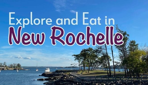 Scenic waterfront view with a calm, blue sky and boats in the distance. Text overlay says, Explore and Eat in New Rochelle, with trees and a rocky shoreline in the foreground.