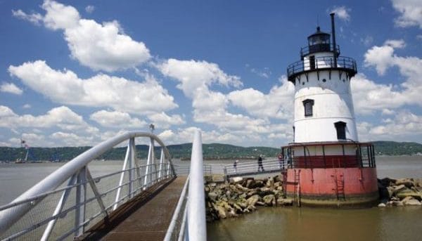 A white and red lighthouse stands on a rocky shore under a partly cloudy sky. A curved pedestrian bridge leads to the lighthouse over the water. Hills are visible in the background.