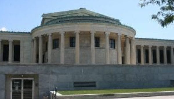 A neoclassical building with a round portico and columns stands under a clear blue sky. The structure is made of stone and features a green, domed roof. Steps lead to an entrance on the left side of the image.