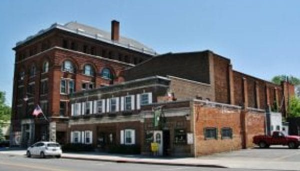 A historic red brick building with arched windows stands on a sunny day. It has a white and brown facade on the lower level. A U.S. flag and a few parked cars are in front.