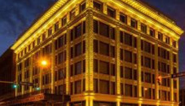 A historic, multi-story building brightly lit with yellow lights outlining its edges against a dark evening sky. Traffic lights and street lamps illuminate the surrounding area, highlighting the buildings architectural details.