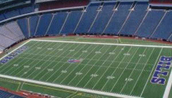 Aerial view of a large, empty football stadium with a green field marked with yard lines. The end zones display the word BILLS in bold letters. Blue and red seating surrounds the field in a semi-circle.