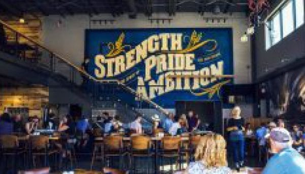 A restaurant interior with people seated at tables. A large mural on the wall reads Strength Pride Ambition in bold letters, surrounded by decorative flourishes. The space has a modern industrial design with high ceilings and exposed beams.