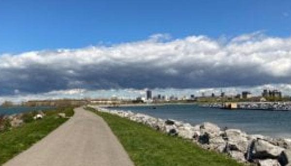 A pathway stretches between a grassy area and a body of water lined with rocks. The skyline of a city is visible under a sky partly covered with dramatic clouds.