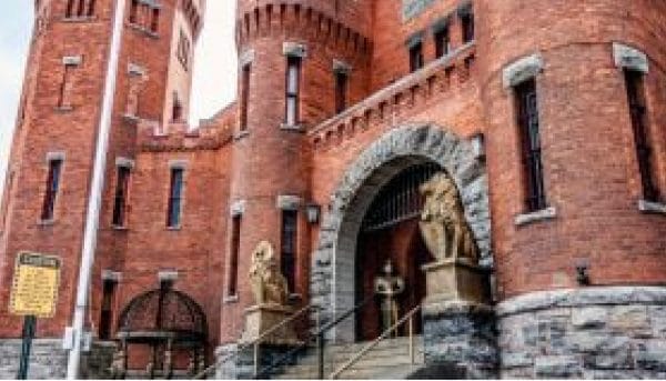 A historic red brick building in central New York, featuring rounded towers and narrow windows, welcomes visitors with an arched entrance flanked by two lion statues. Stone steps lead up to the entrance, with a road sign visible to the left.