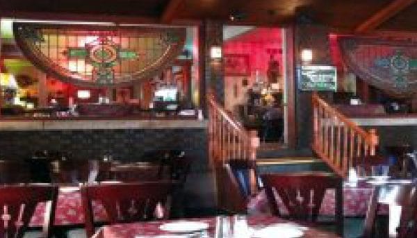 A cozy restaurant interior with wooden furniture, decorative stained glass panels, and dim lighting evokes the charm of a hidden gem near Niagara Falls. Several tables are set, and stairs lead to an upper level with more seating, creating a warm and inviting atmosphere.