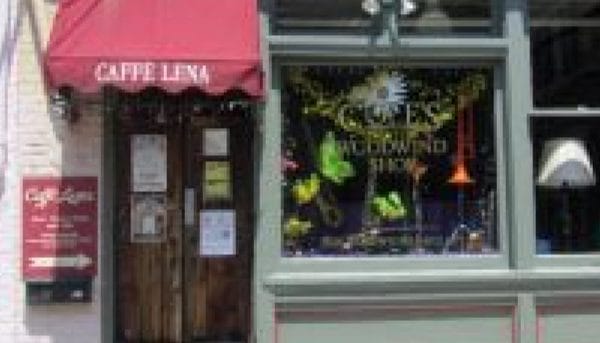The exterior of Caffe Lena in Saratoga Springs showcases a red awning proudly displaying its name. The window features decorative elements like hanging lights and vibrant artwork, while the door to the left is adorned with various posted signs. Inside, a vintage lamp adds charm to this iconic locale.