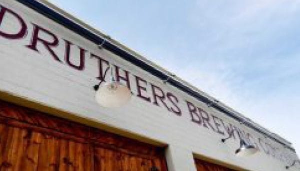 Low-angle view of Druthers Brewing Companys exterior in Saratoga Springs, featuring white walls and wooden doors. Large letters spell out the name, while two outdoor lights adorn the entrance. The sky above is partly cloudy, adding a touch of drama to this charming local gem.