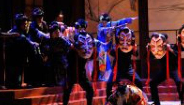 Performers in colorful costumes and masks take the stage during a theatrical performance in Saratoga Springs. Dramatic lighting highlights their vibrant attire and expressive gestures, creating a striking contrast against the dim background.